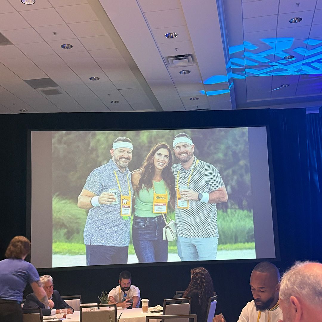 Conference room with a large screen showing three people posing with drinks, audience seated below.