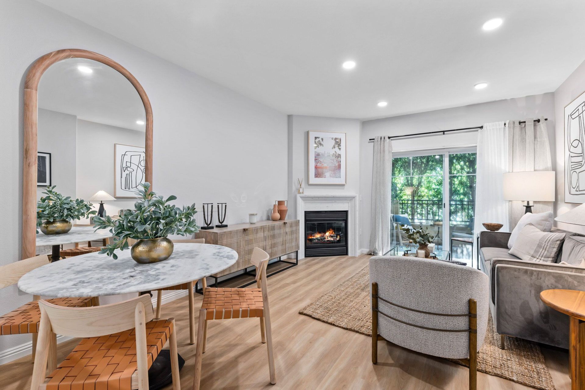 Bright modern living room and dining area with fireplace, large mirror, gray sofa, and sliding doors to greenery