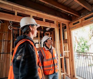 Two construction workers in hard hats and orange vests inspecting a framed building interior
