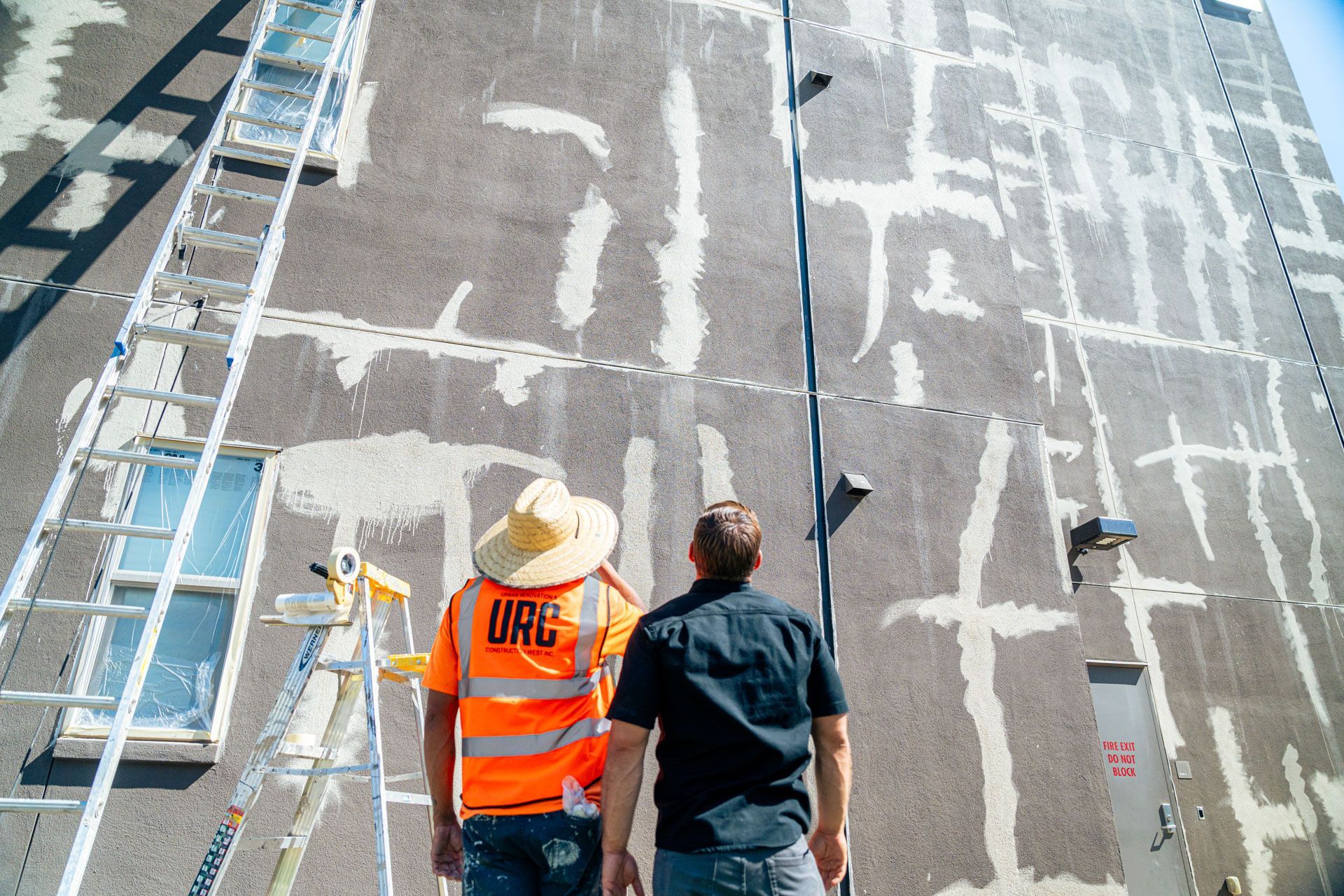 Two workers in safety vests inspect a tall building facade with scaffolding and mural markings