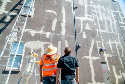 Two workers in safety vests inspect a tall building facade with scaffolding and mural markings