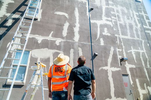Two workers inspect a tall building facade with white patch marks and a blue lift scaffold.