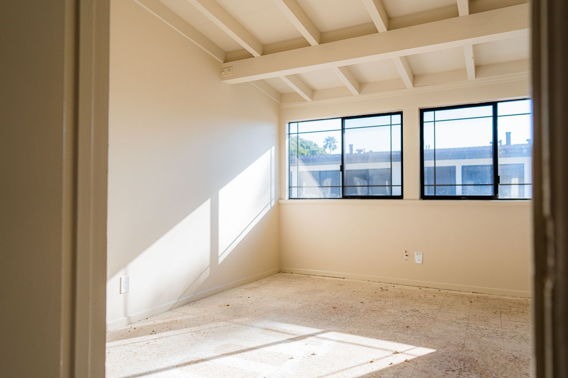 Empty bright room with beige walls, exposed beams, and large windows casting sunlight on the floor.
