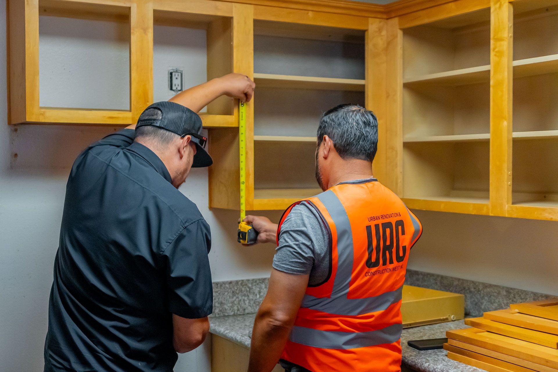 Two workers installing a kitchen cabinet in a room with yellow wood cabinets and a gray backsplash