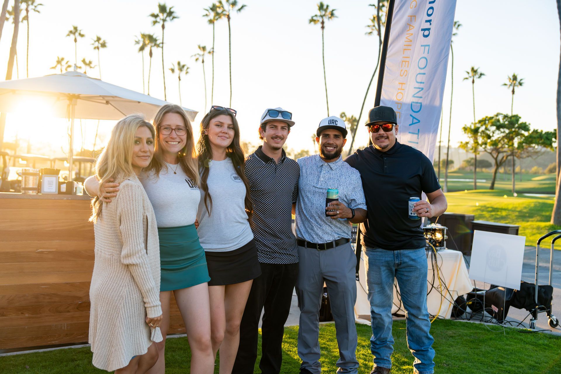 Five friends smiling outdoors at sunset, standing near a grassy event area with palm trees and a stage.