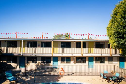 Motel-style building with balconies, red bunting, and a poolside patio under a clear blue sky