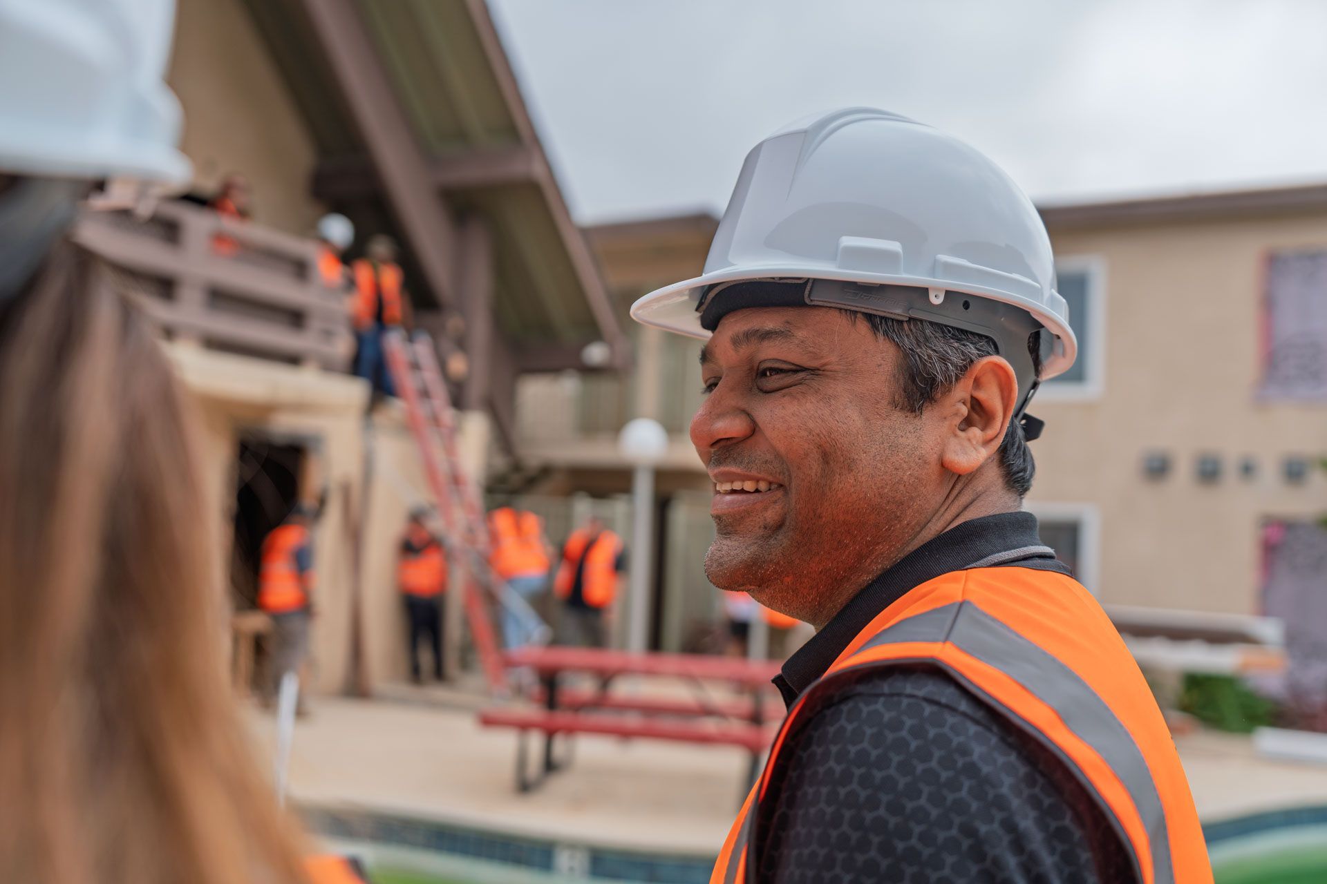 Worker in gray hard hat and orange safety vest at a construction site, looking over his shoulder.