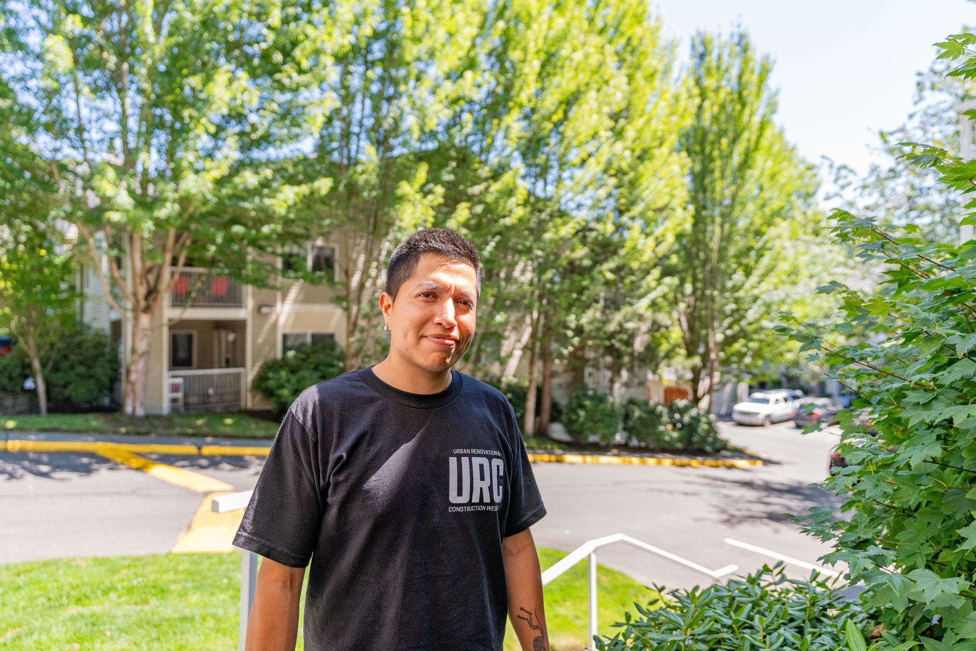 Man standing outdoors in a black shirt on a sunny tree-lined street near apartments