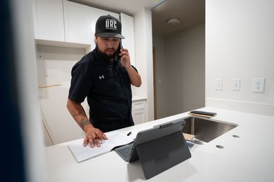 Person on phone in a modern kitchen, leaning on a white counter beside a tablet and open sink.