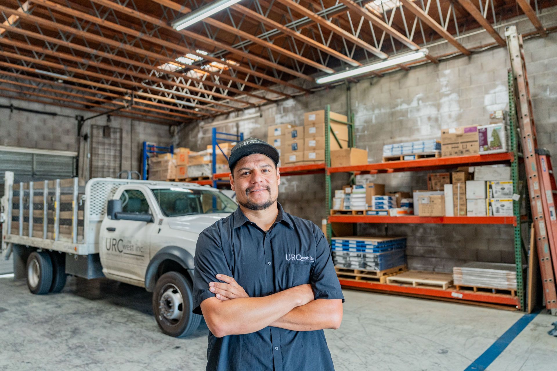 Warehouse worker standing with arms crossed beside a flatbed truck and storage shelves in a warehouse.