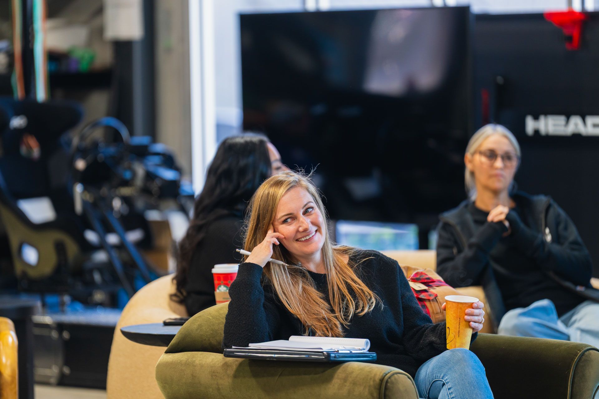 Woman smiling on a couch in a casual indoor lounge, with others seated in the background.