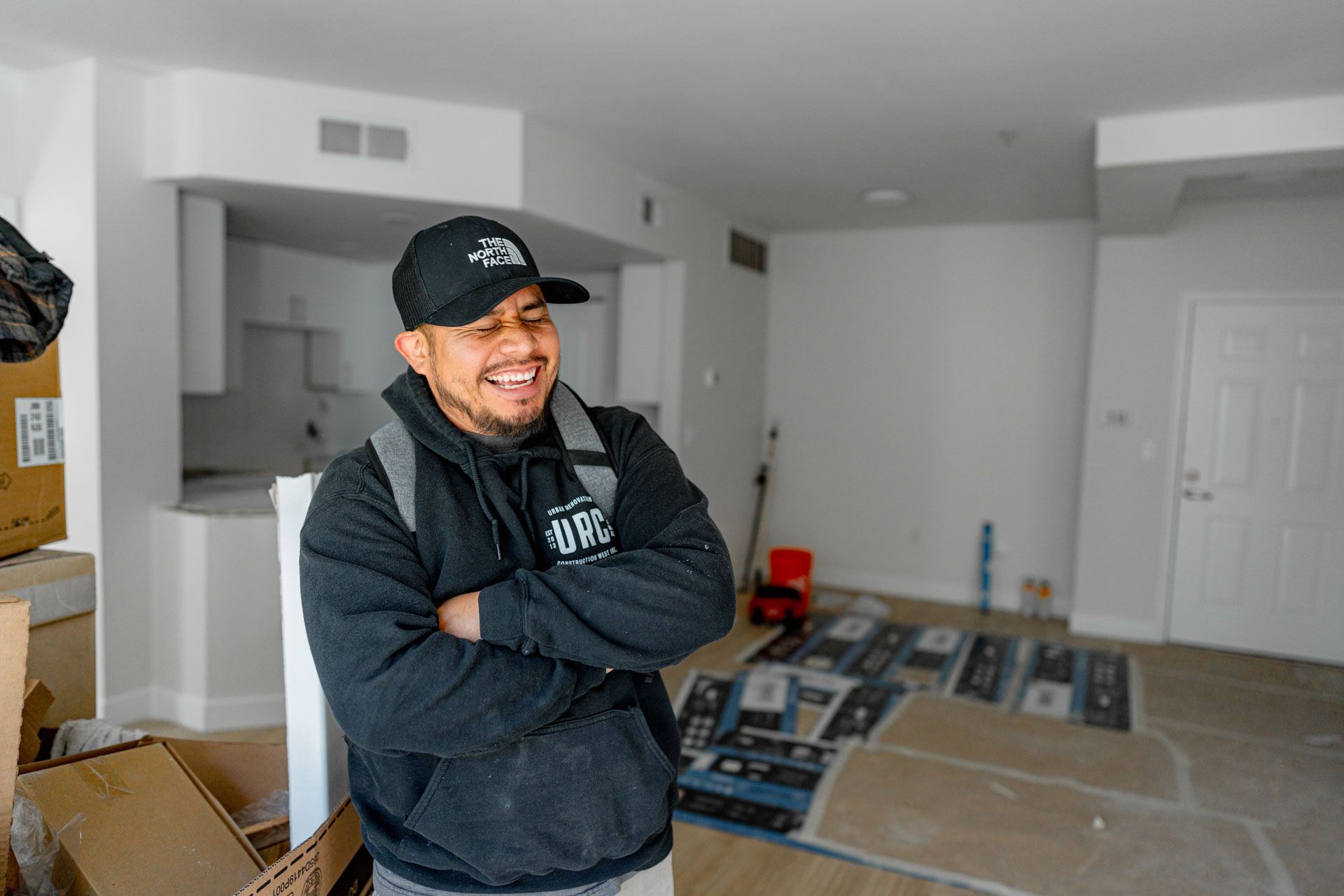 Smiling man with arms crossed in an unfinished room with tools, boxes, and a ladder on the floor
