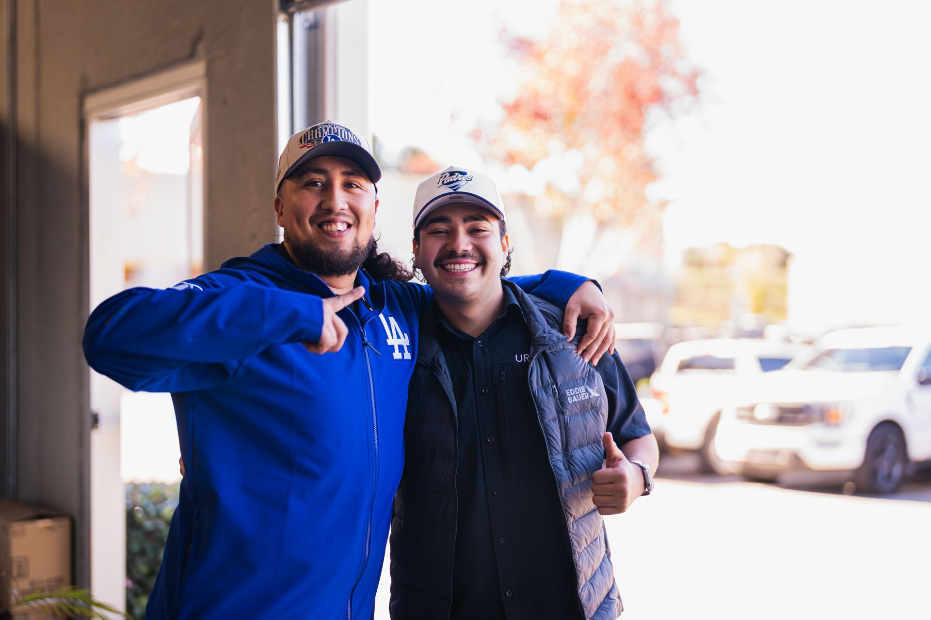 Two smiling men in blue jackets posing together at a doorway, one giving a thumbs-up
