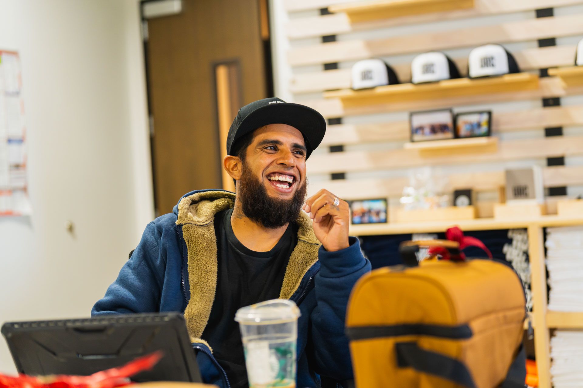 Smiling man in a cap sitting at a cafe table with a laptop and iced drink, blurred shelves in background