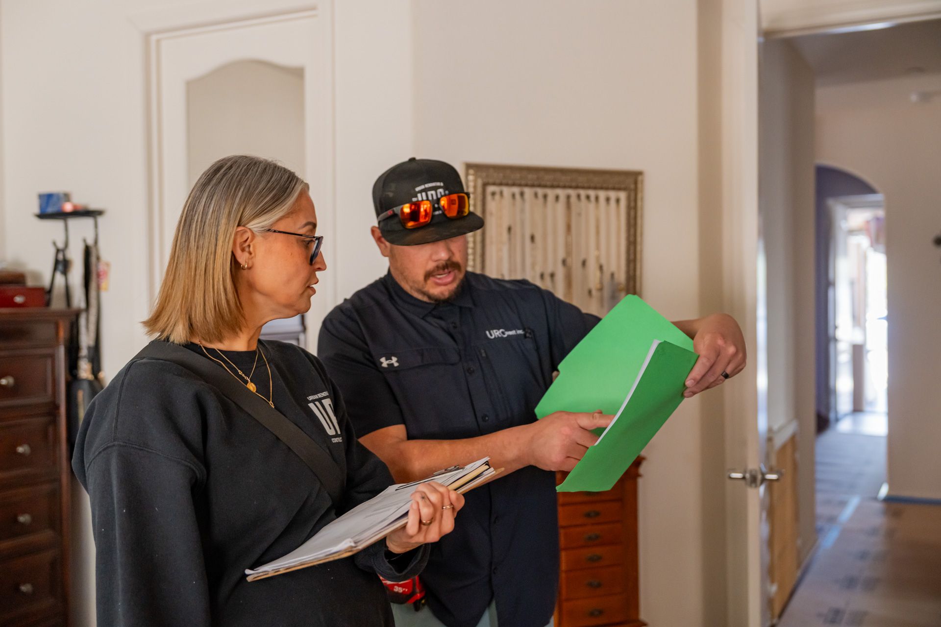 Two people review paperwork in a hallway, one holding a green folder and the other a clipboard.