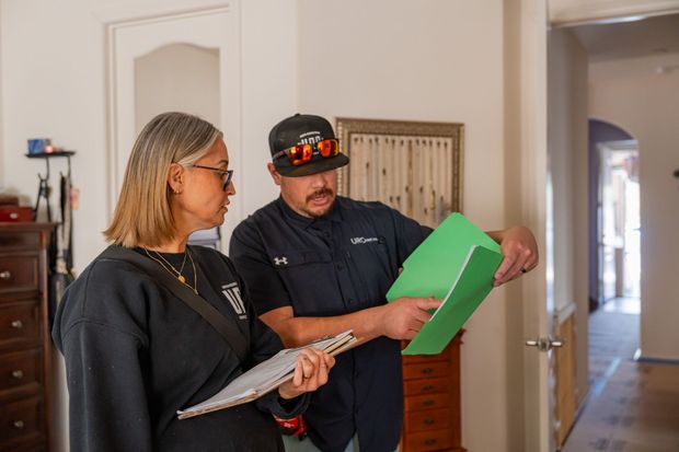 Two people review paperwork in a hallway, one holding a green folder and the other a clipboard.