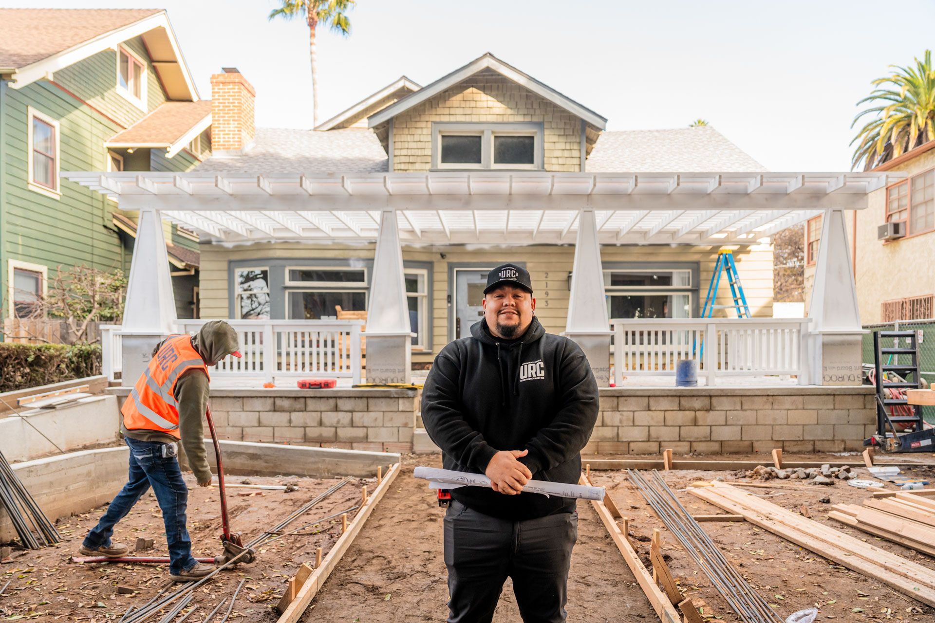 Two workers stand in front of a house under construction with exposed framing and bare ground.