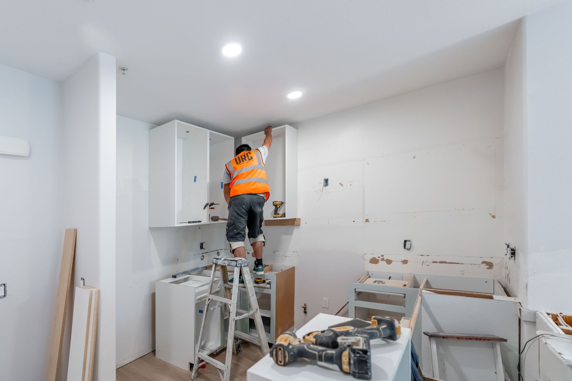 Worker on a ladder installing cabinets in a white kitchen under renovation.