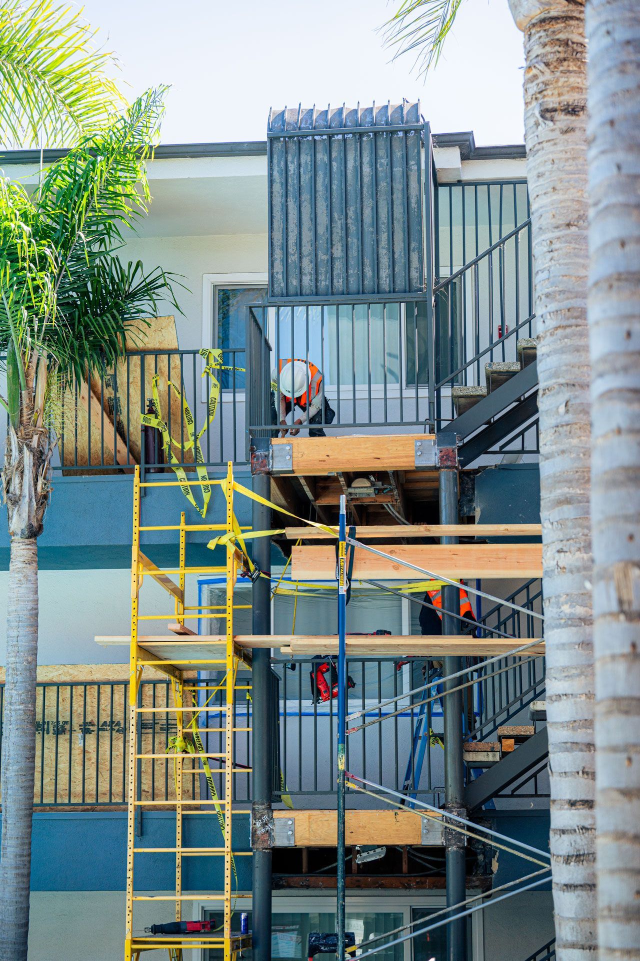 Scaffold and stairs outside a building with workers and yellow safety rails, framed by palm trees