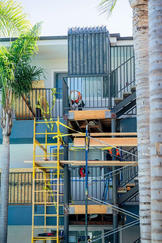 Workers on scaffolding beside a beige building, with palm trees in the foreground