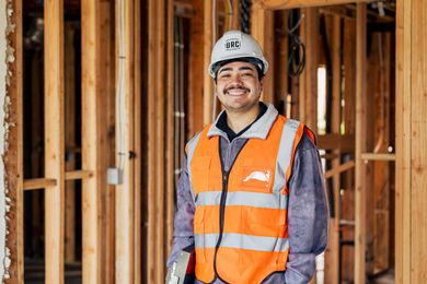 Construction worker in a hard hat and orange safety vest smiling inside a wooden-framed building site
