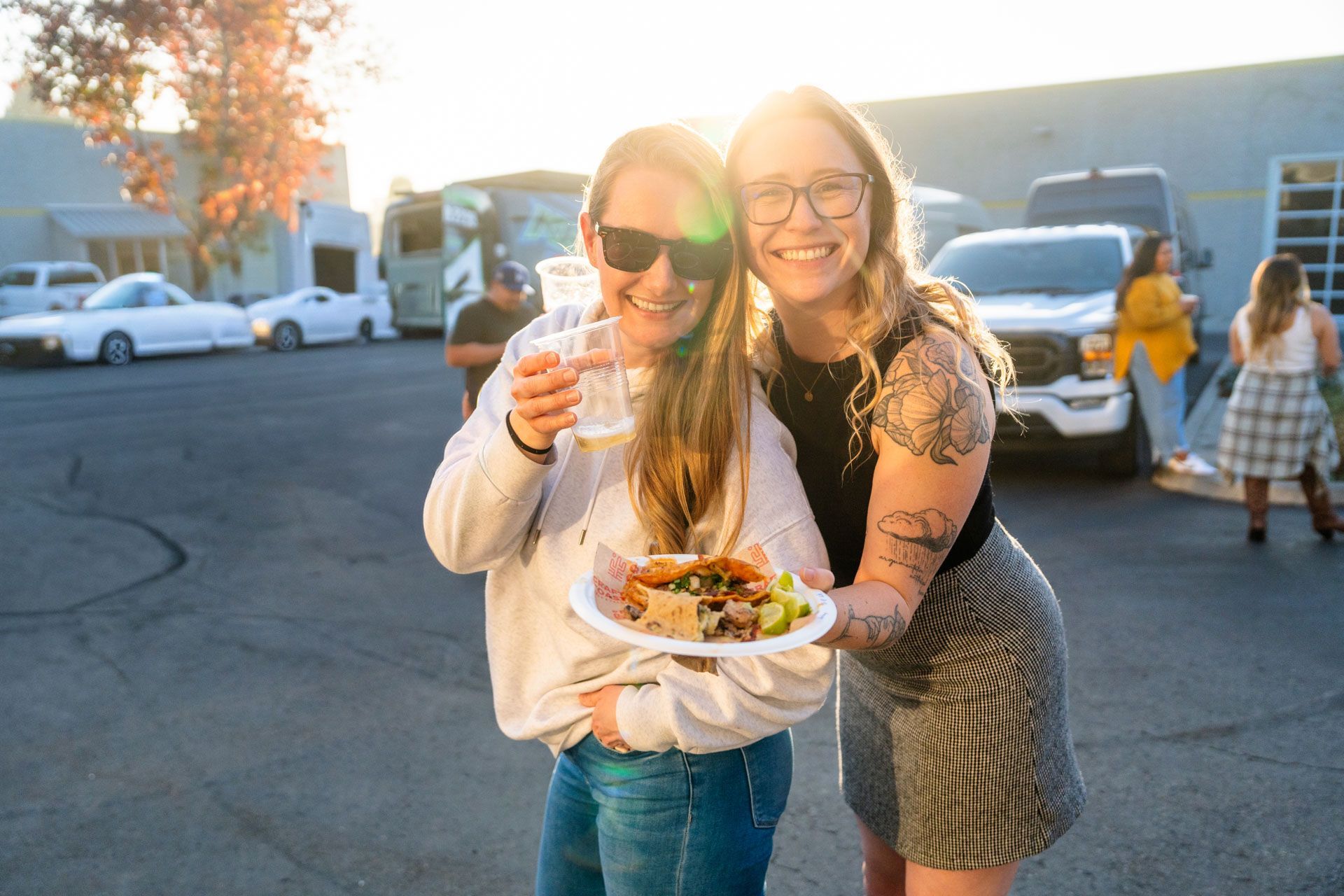 Two people smiling and holding drinks and a plate of food outdoors at sunset
