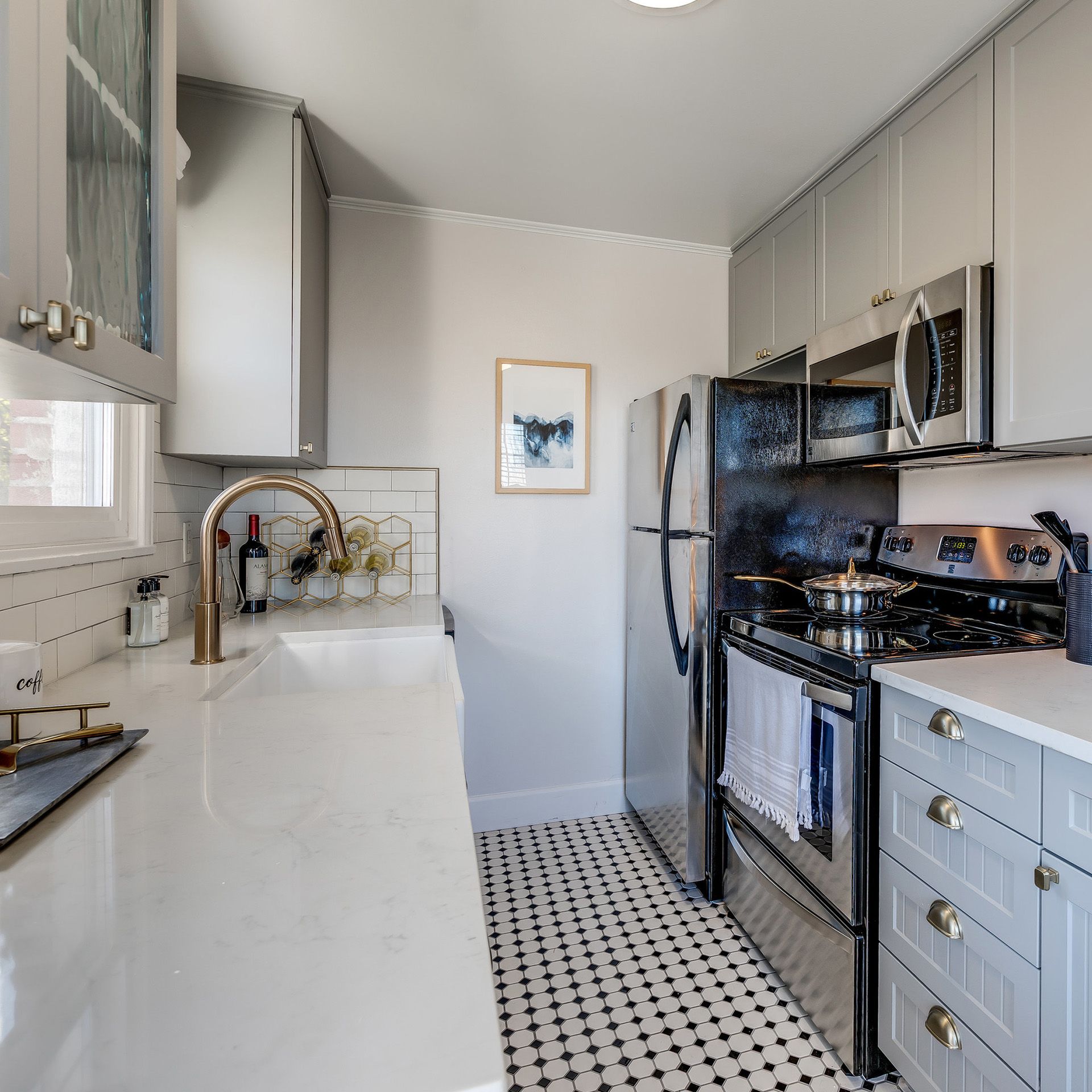 Narrow modern kitchen with white cabinets, black stove, stainless fridge, and black-and-white floor tiles