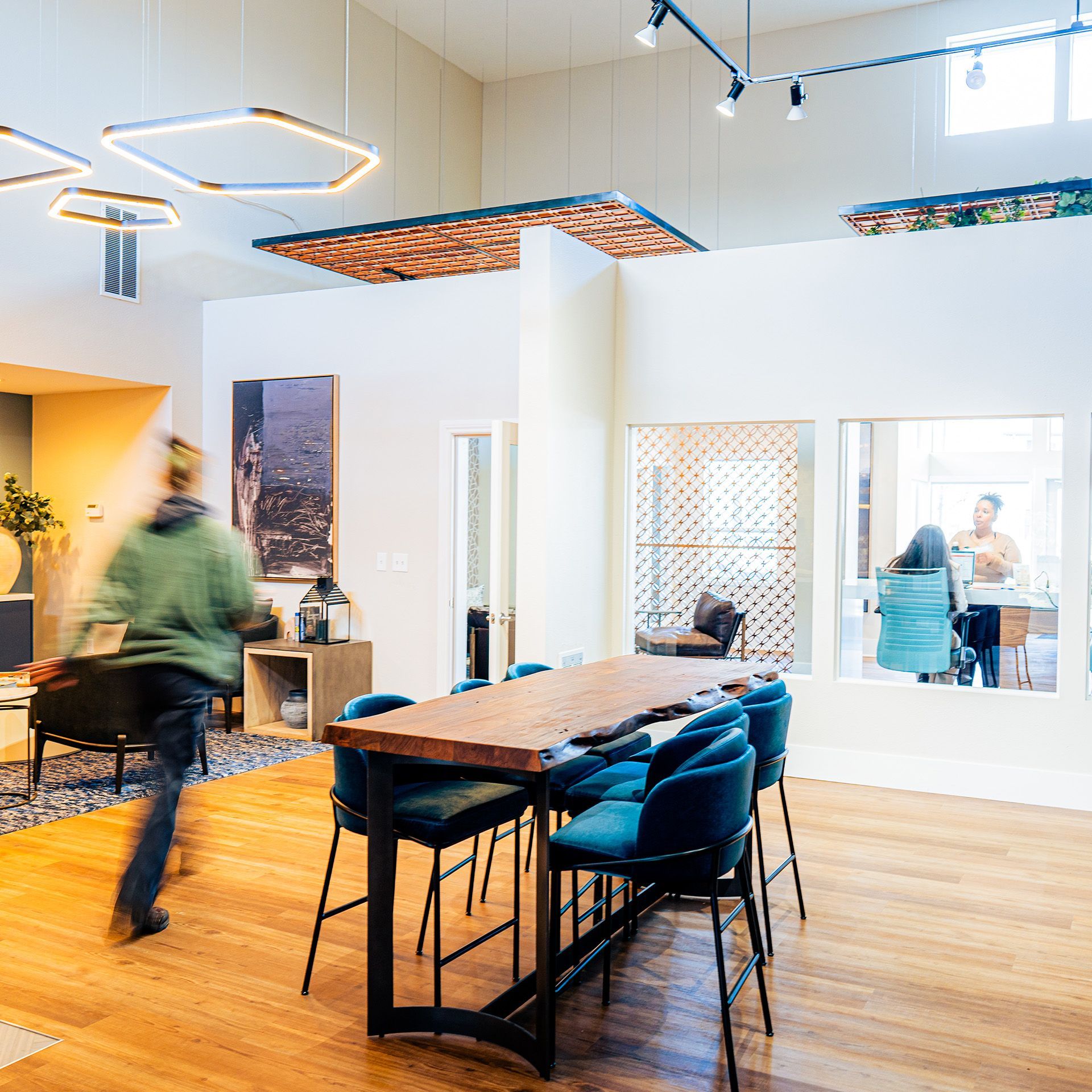 Modern office lounge with wooden table, blue chairs, and people moving in a bright open workspace