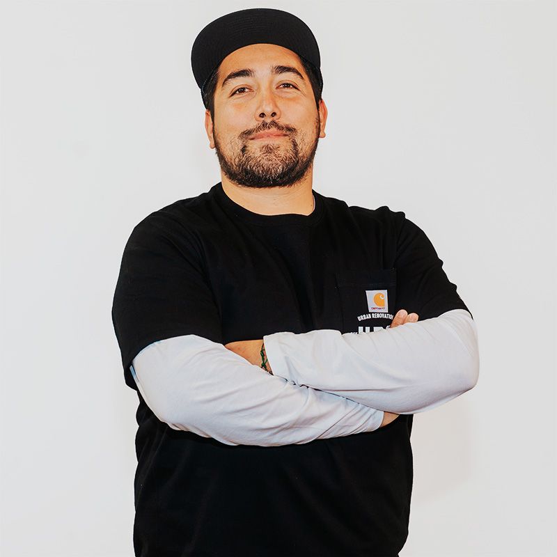 Man standing with arms crossed, wearing a black cap and black shirt over long white sleeves, against a white background