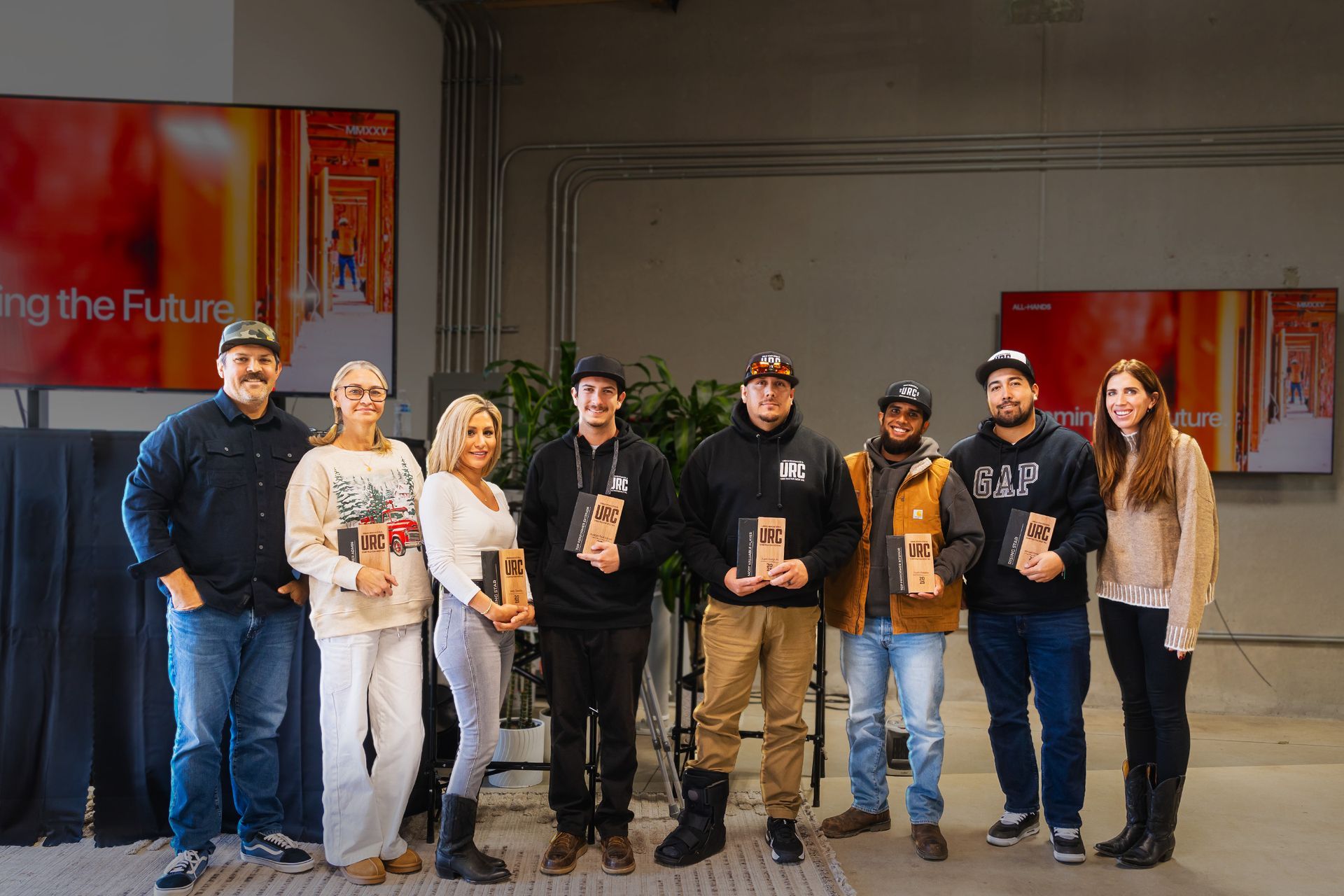 Eight people standing in a row indoors, holding awards or plaques in front of a gray wall and banners.