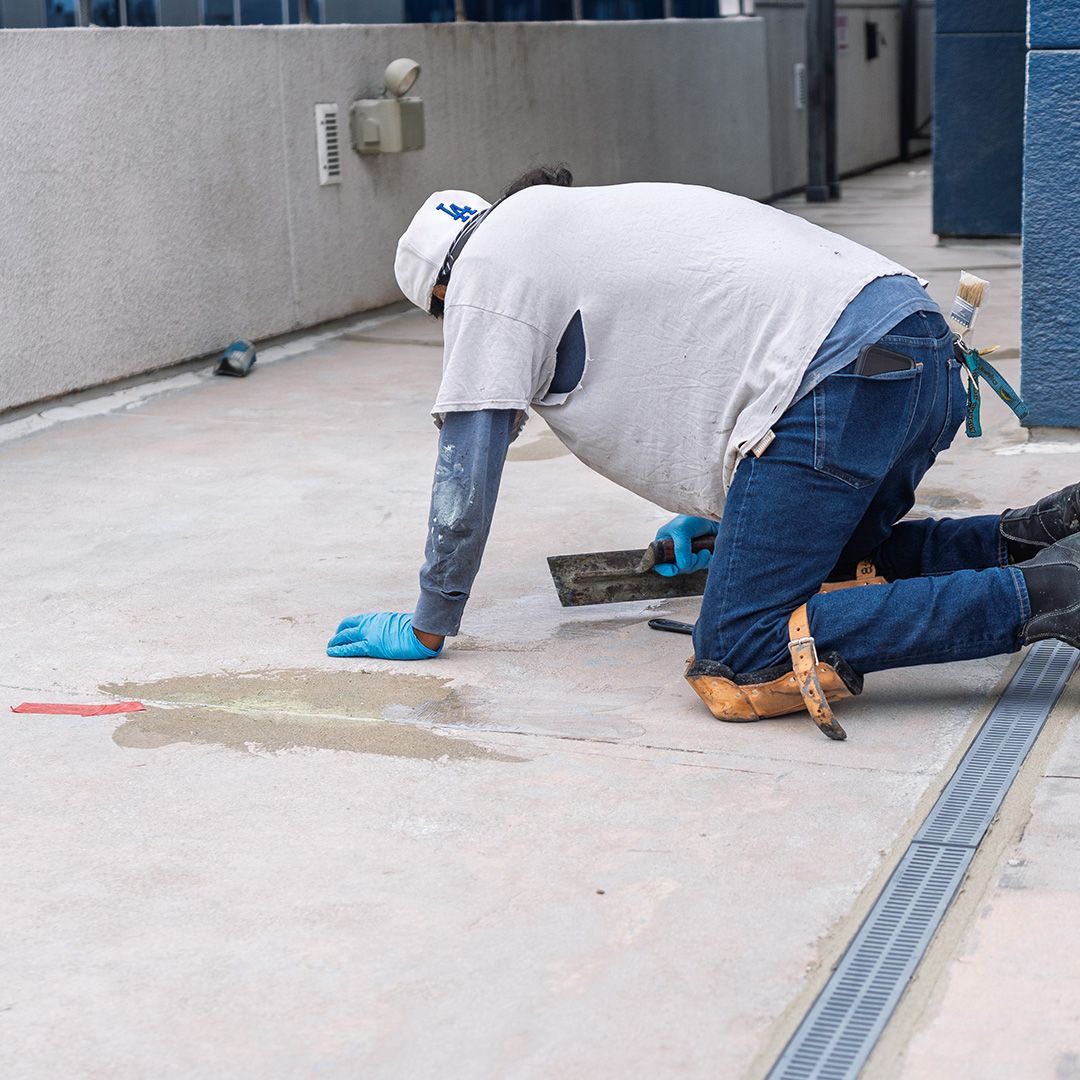 Person on knees repairs concrete floor, wearing gloves, knee pads, and hat. Gray floor, blue wall.
