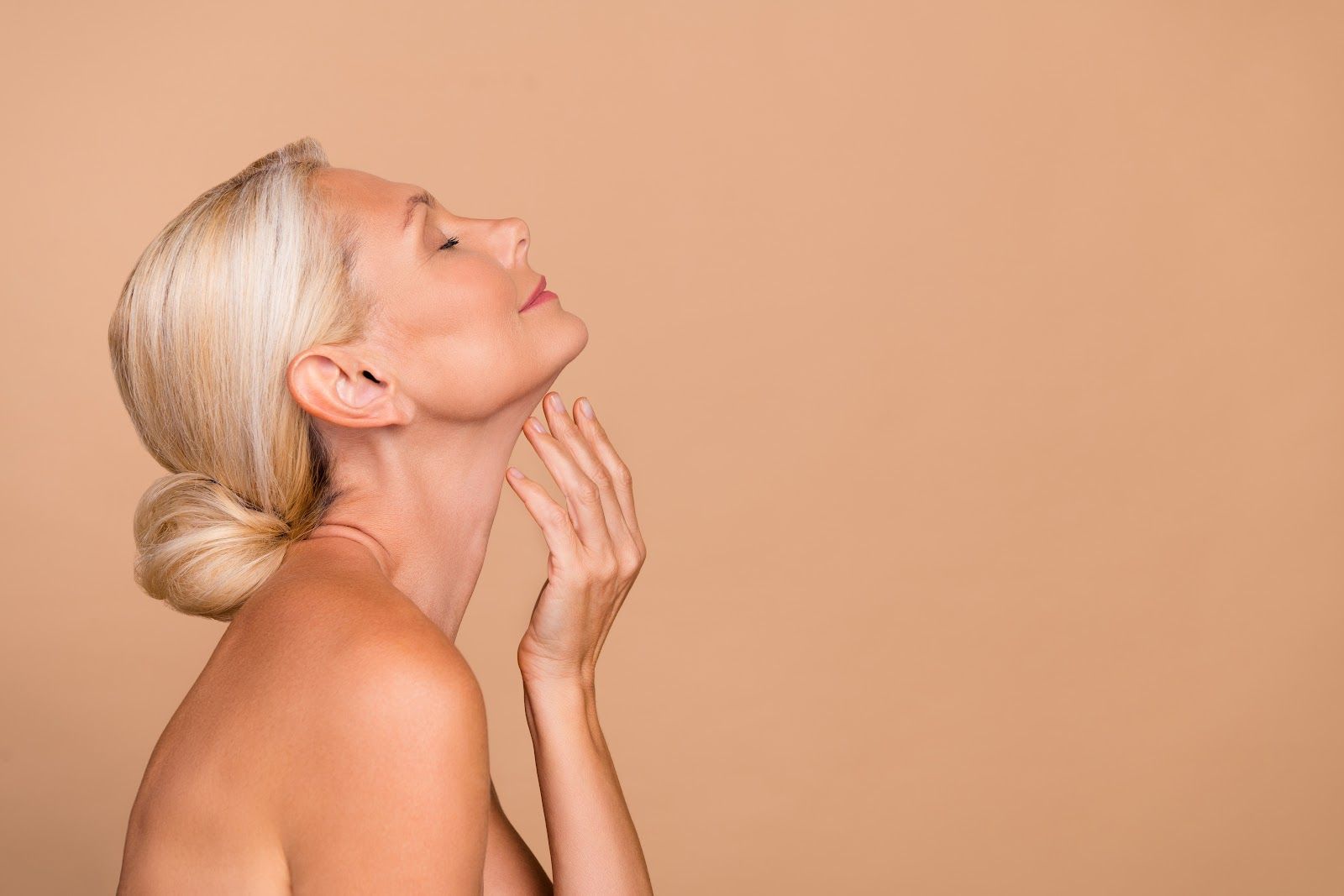 Blonde woman in profile applying skincare to her neck against a peach background