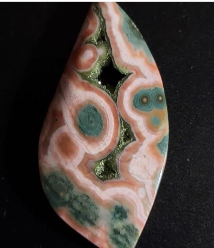 Polished teardrop-shaped gemstone with concentric rings of peach, green, and white patterns, and a dark, central cavity.