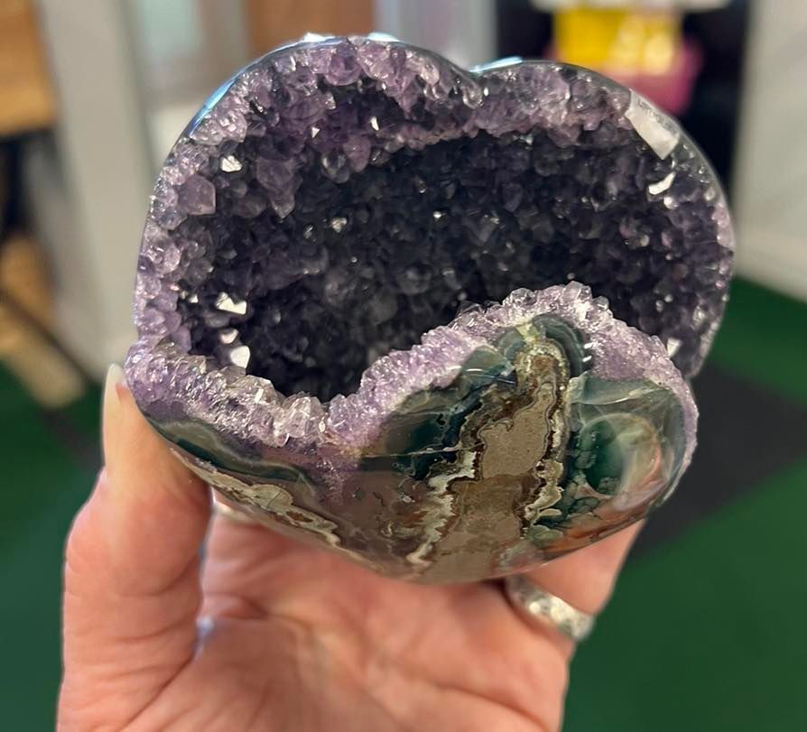Hand holding heart-shaped amethyst geode, purple crystals inside, green and brown exterior.