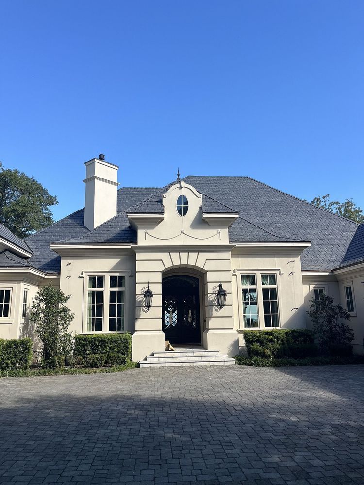 Cream-colored house with a dark roof, arched front entry, and paved driveway under a blue sky