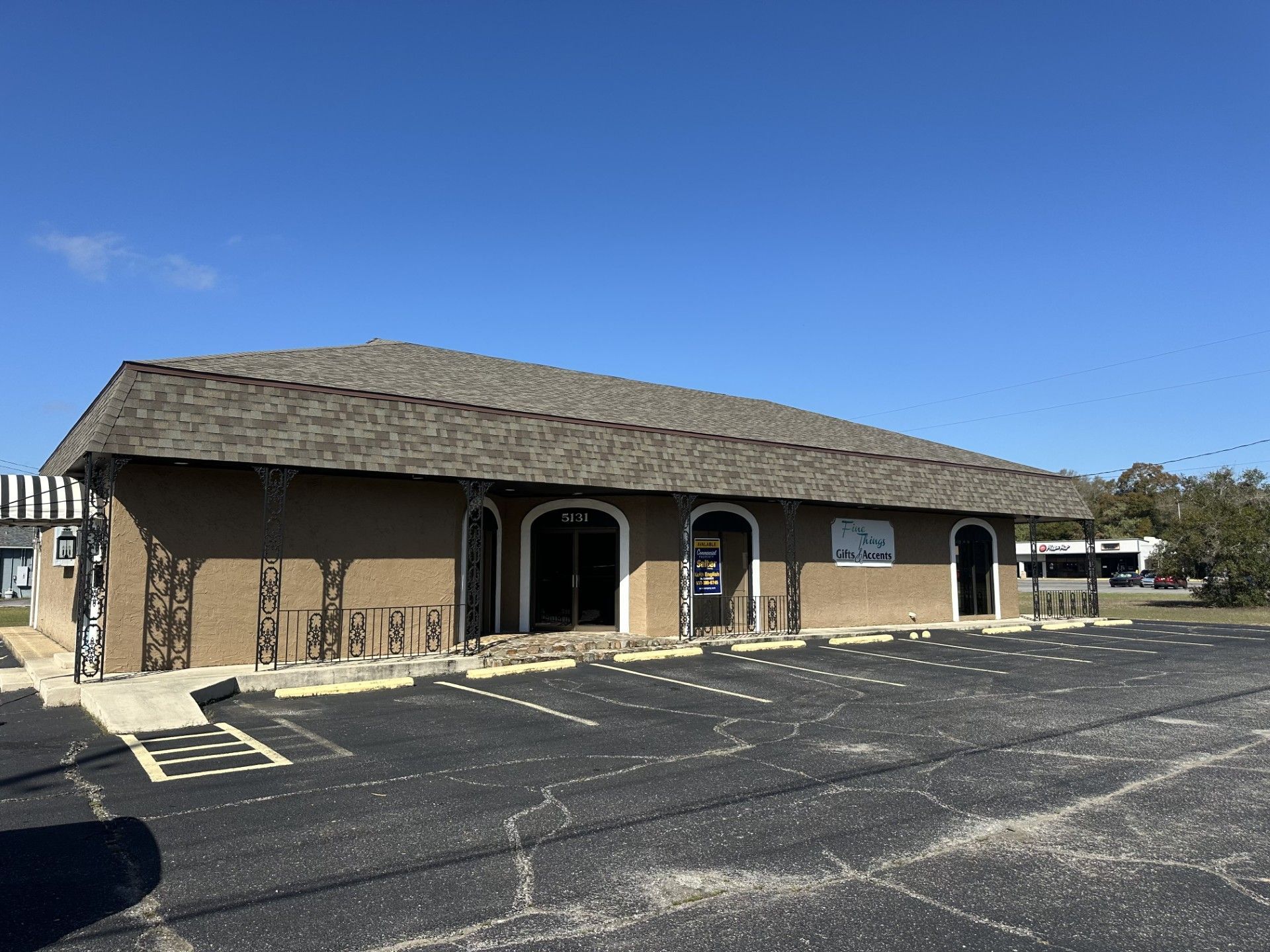 Single-story tan building with arched windows and a dark roof beside an empty parking lot under a clear blue sky