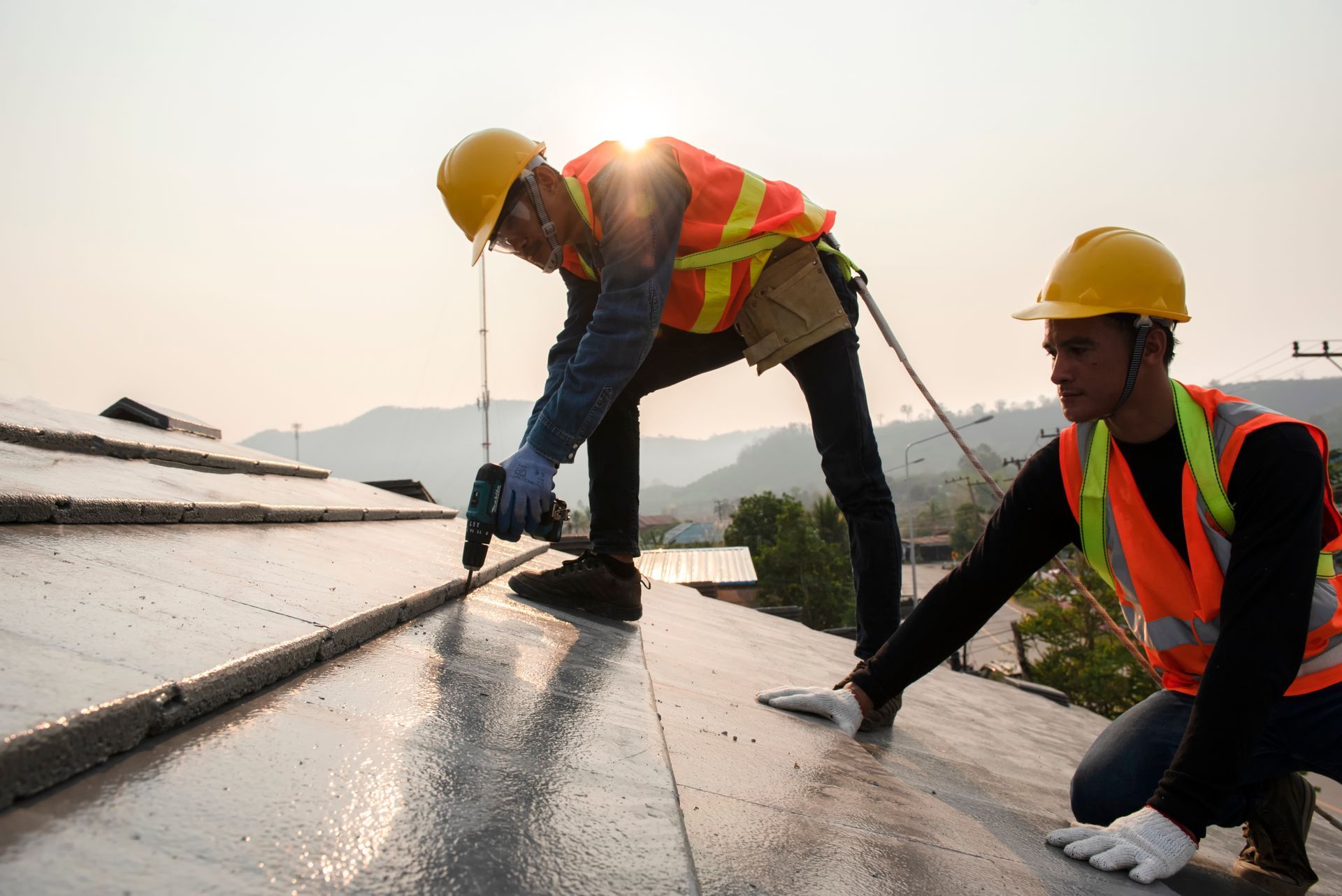 Two workers in hard hats and safety vests inspecting a wet rooftop with tools at sunset