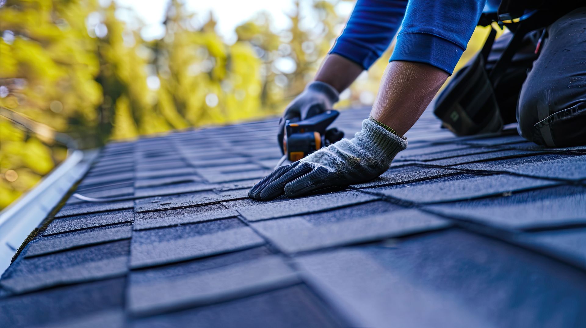 Residential roofing services worker repairing shingle roof with nail gun and safety gloves.