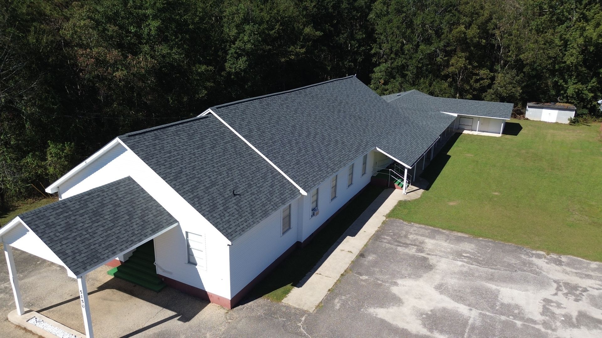 Aerial view of a long white building with dark roof beside a grassy field and paved lot.