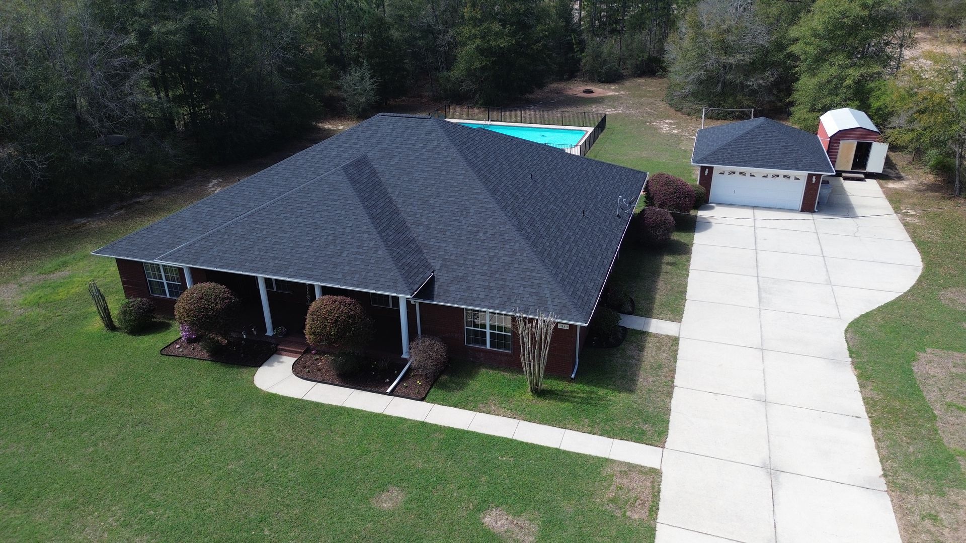 Aerial view of a brick house with a dark roof, long driveway, pool, and detached garage on a grassy lot