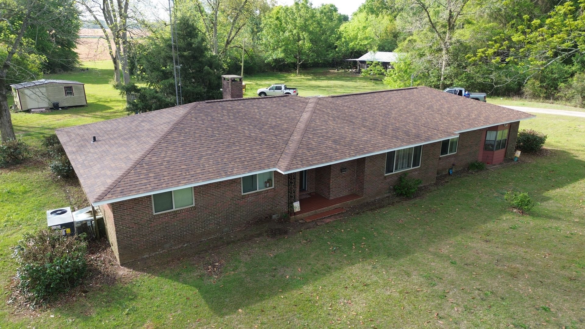 Aerial view of a single-story brick house with a brown roof, surrounded by a grassy yard and trees.