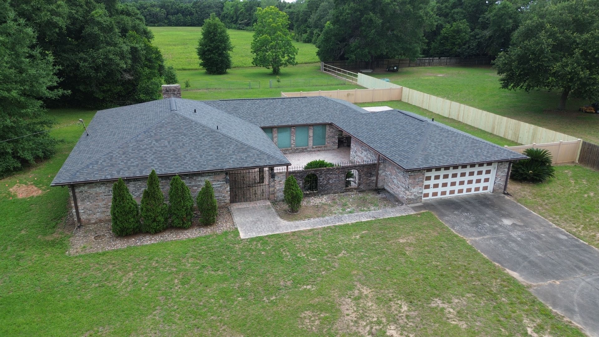 Aerial view of a large stone house with a dark roof, driveway, and green lawn surrounded by trees.
