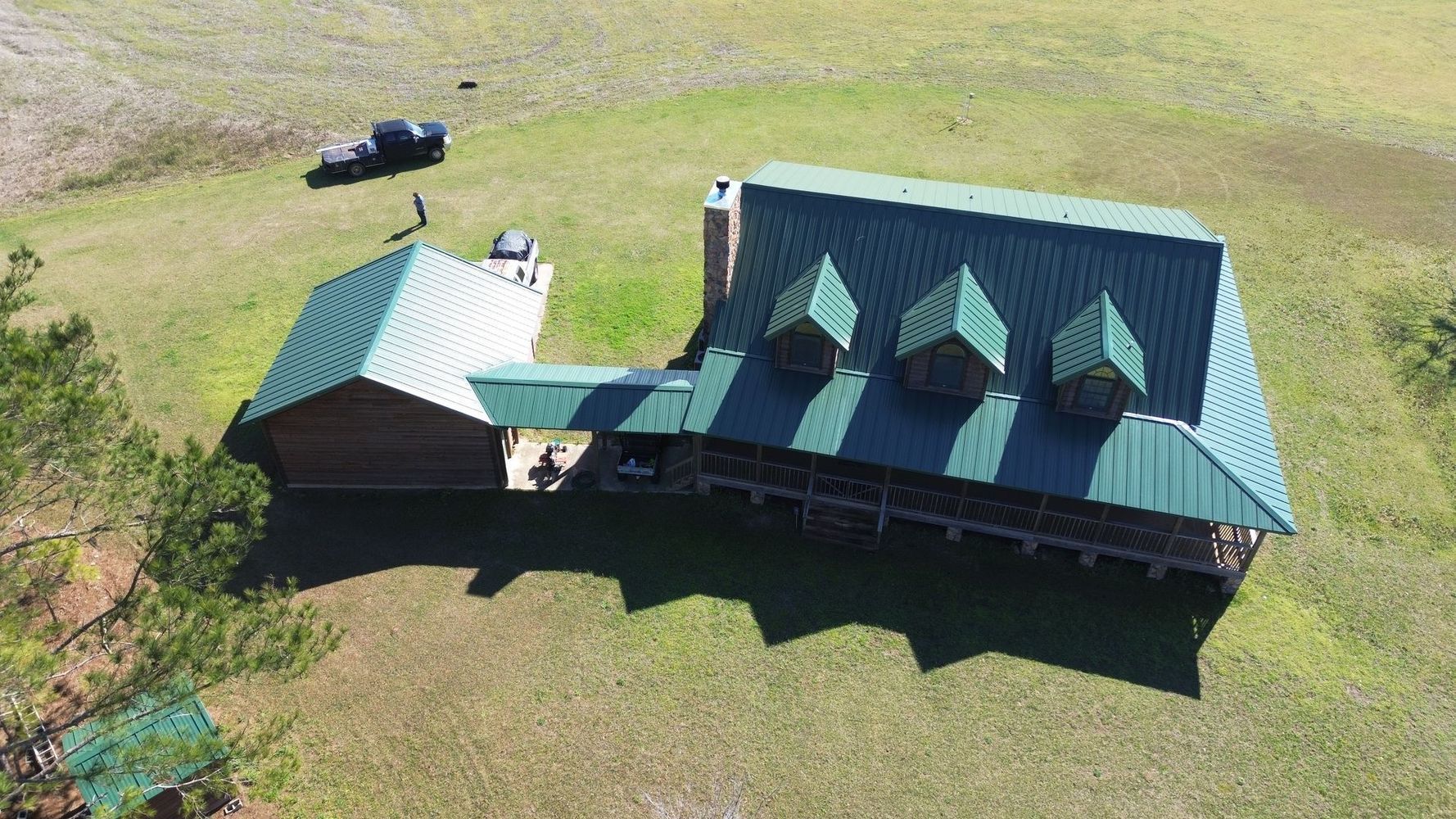 Aerial view of a green-roofed house and barn on a grassy rural property, with a black vehicle nearby
