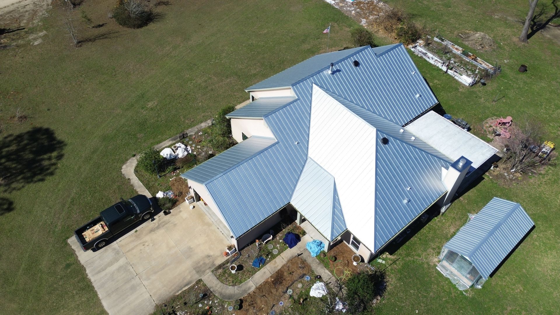 Aerial view of a white house with a blue metal roof and driveway in a rural area