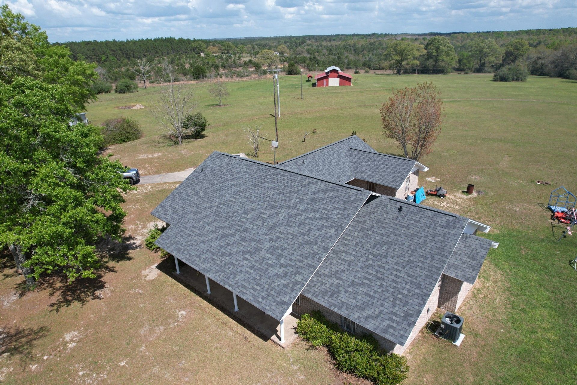 Aerial view of a gray-roofed house with a yard, trees, and a red barn in the distance.