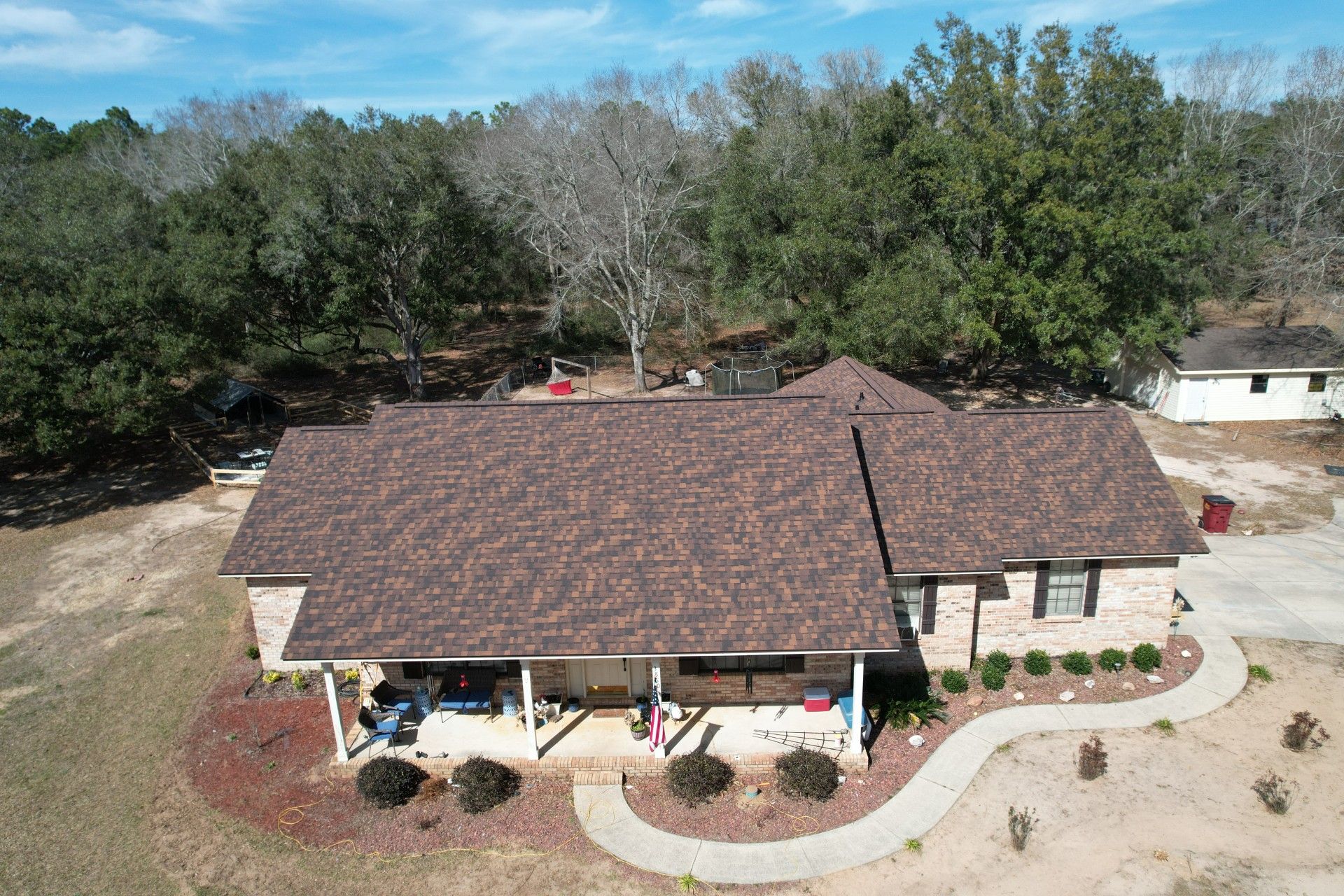 Aerial view of a single-story house with a brown roof, front porch, and curved walkway surrounded by trees.