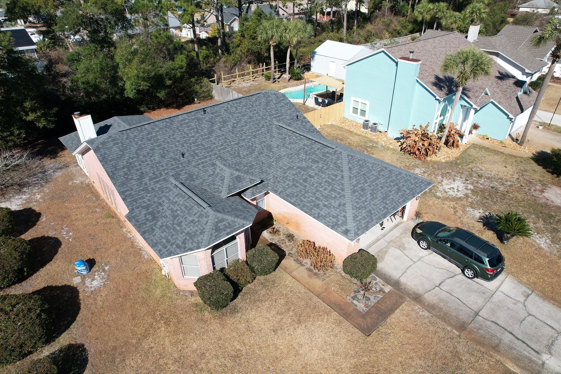 Aerial view of a single-story house with a gray roof, driveway, and a parked car in a suburban neighborhood.