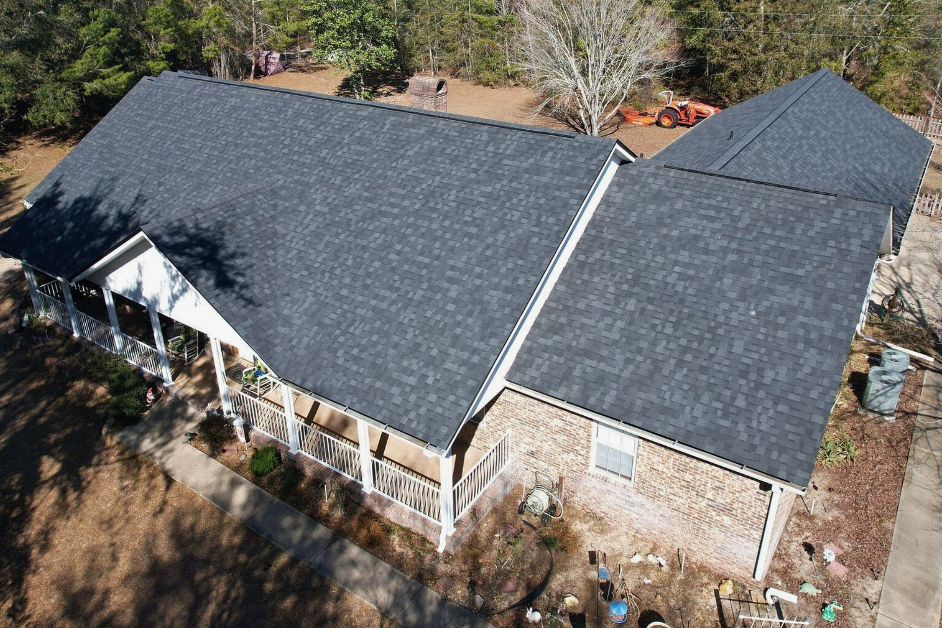 Aerial view of a house with a dark gray shingle roof and brick exterior surrounded by trees.