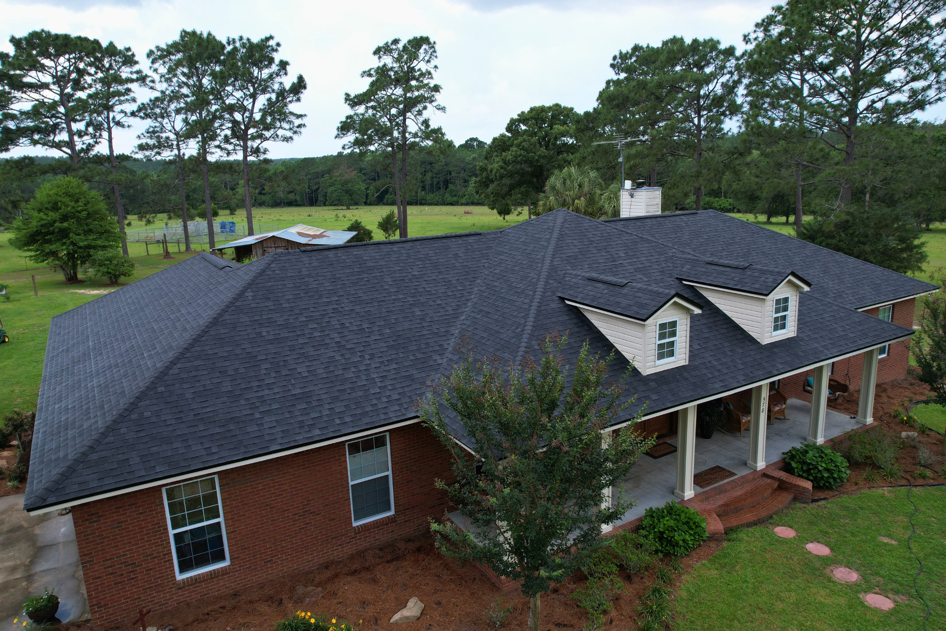 Aerial view of a red brick house with a dark roof, white porch, and surrounding trees and lawn