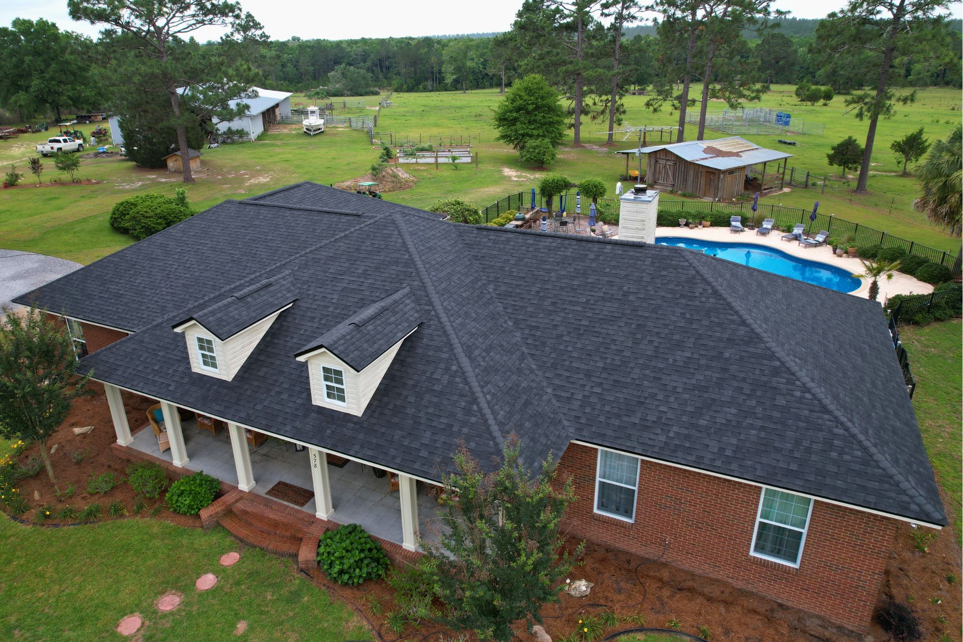 Aerial view of a large brick house with a dark roof beside a backyard pool and green lawn.